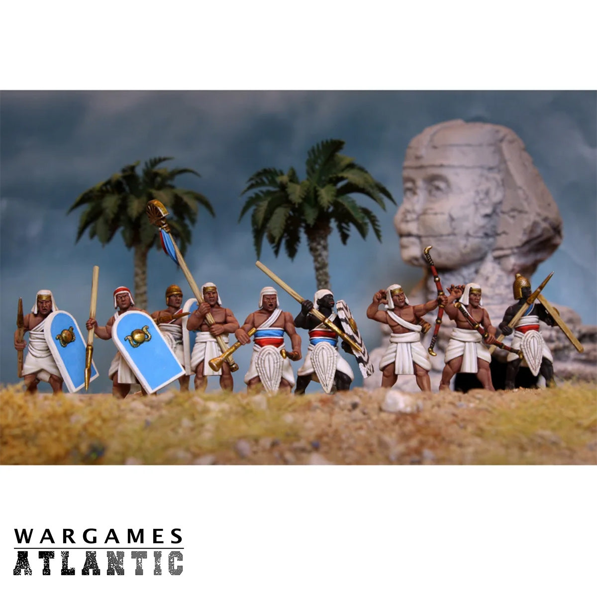 A set of Wargames Atlantic ancient Egyptian soldier miniatures arranged in a diorama with palm trees, a large stone head statue, and a cloudy sky backdrop. The bottom of the image features a white banner with the 'WARGAMES ATLANTIC' logo.