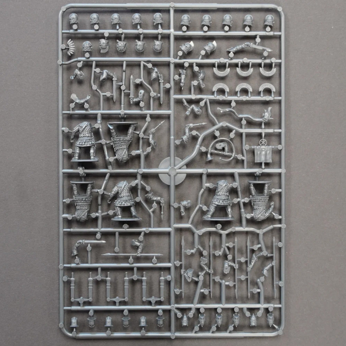 An unpainted gray plastic sprue containing various Roman miniature figurines, weapons, and accessories for model building, photographed flat against a neutral gray background.