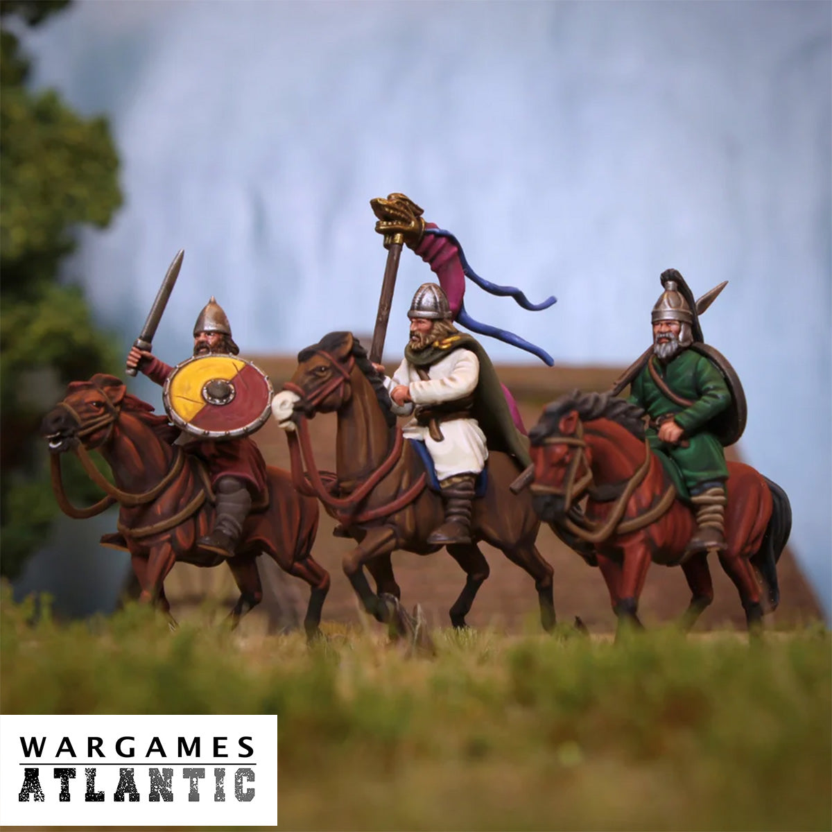 Three painted wargaming miniature riders on horseback, with helmets, shields, swords, and a banner, set in a grassy tabletop terrain with a blurred outdoor-style background. A Wargames Atlantic logo appears in the lower left corner.